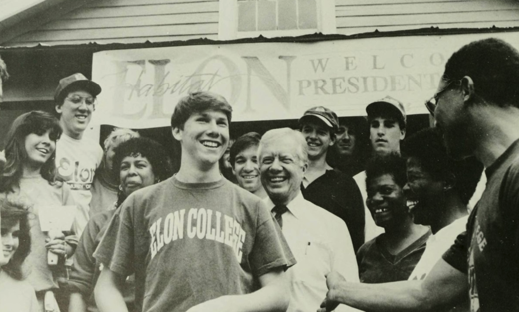 A black and white photo showing a group of smiling people gathered around a student wearing an Elon College t-shirt, shaking hands with former U.S. President Jimmy Carter under a banner that reads "Welcome the President Carter.""