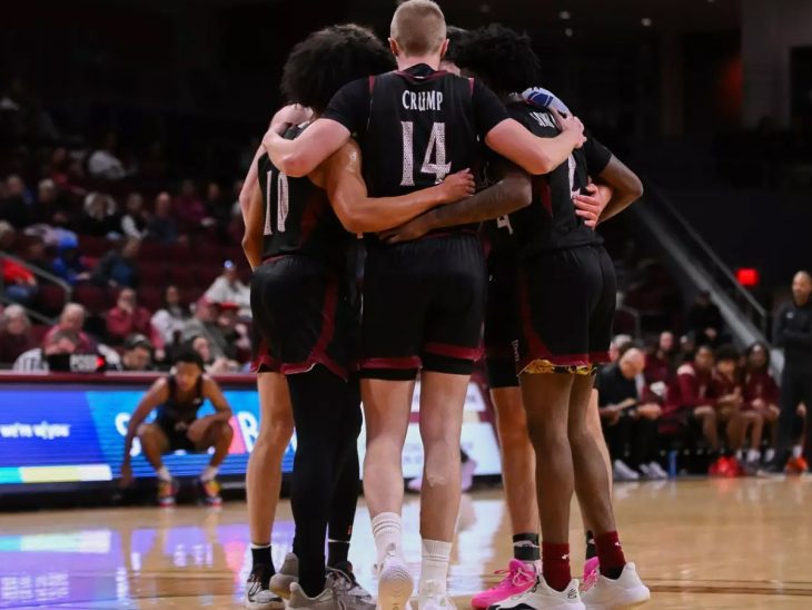 Five basketball players in maroon and black uniforms huddle on the court, their arms around each other. The name "CRUMP" is visible on the jersey of one player.