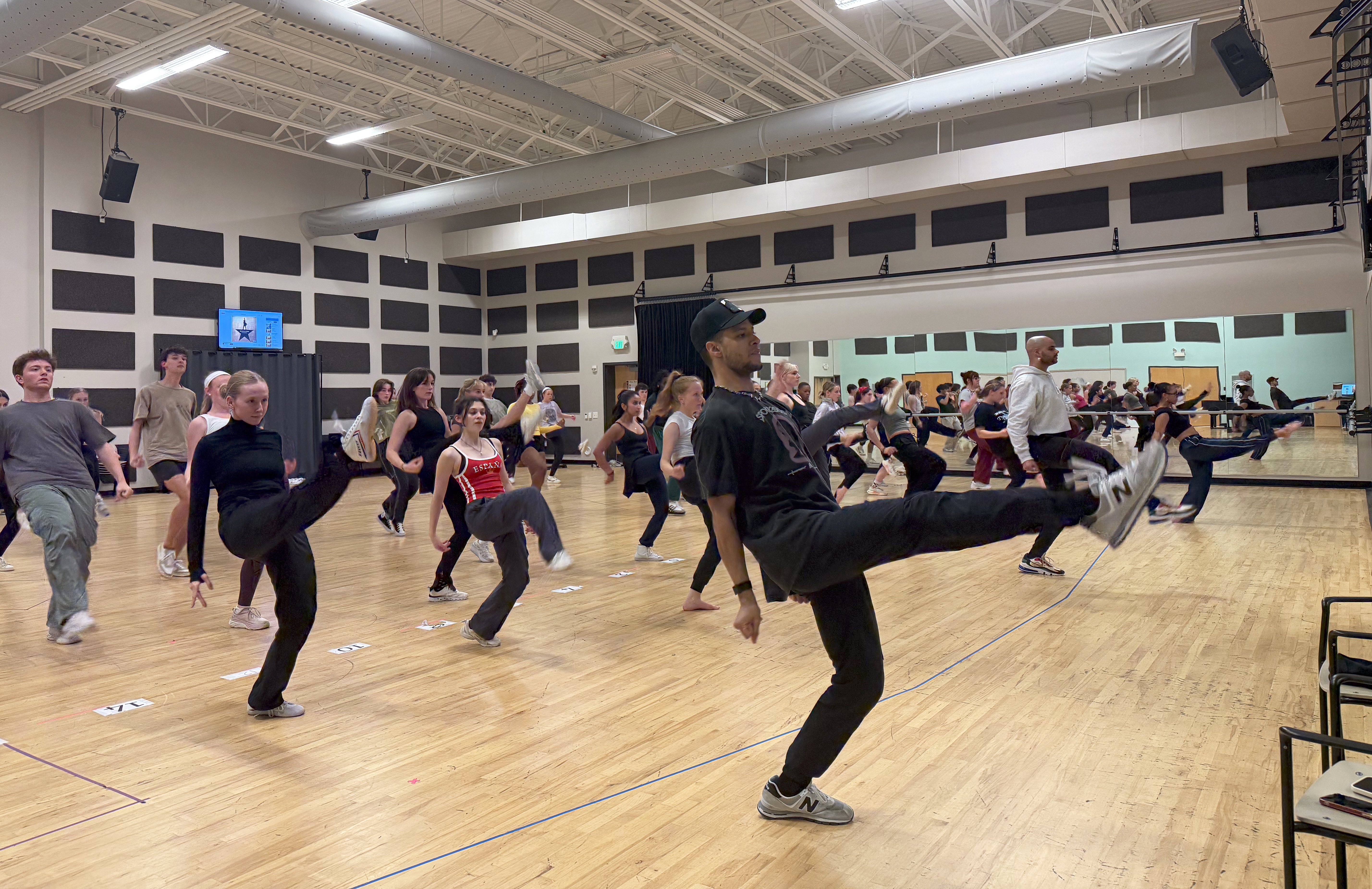 A large dance rehearsal room with about 30 people kicking their right legs up in a dance routine