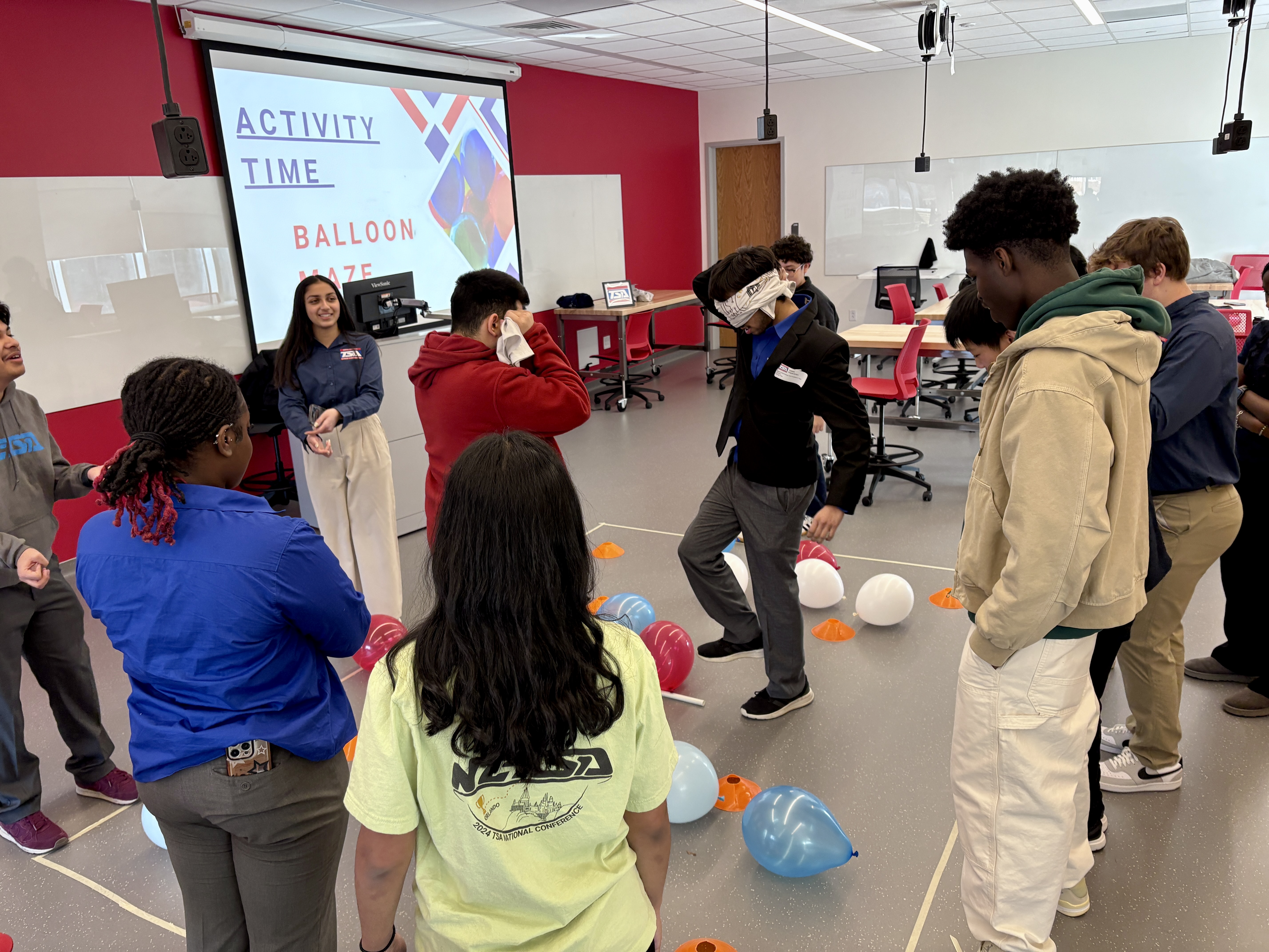 Two students with bandaged eyes cross a maze of balloons and orange cones while the teammates encourage them