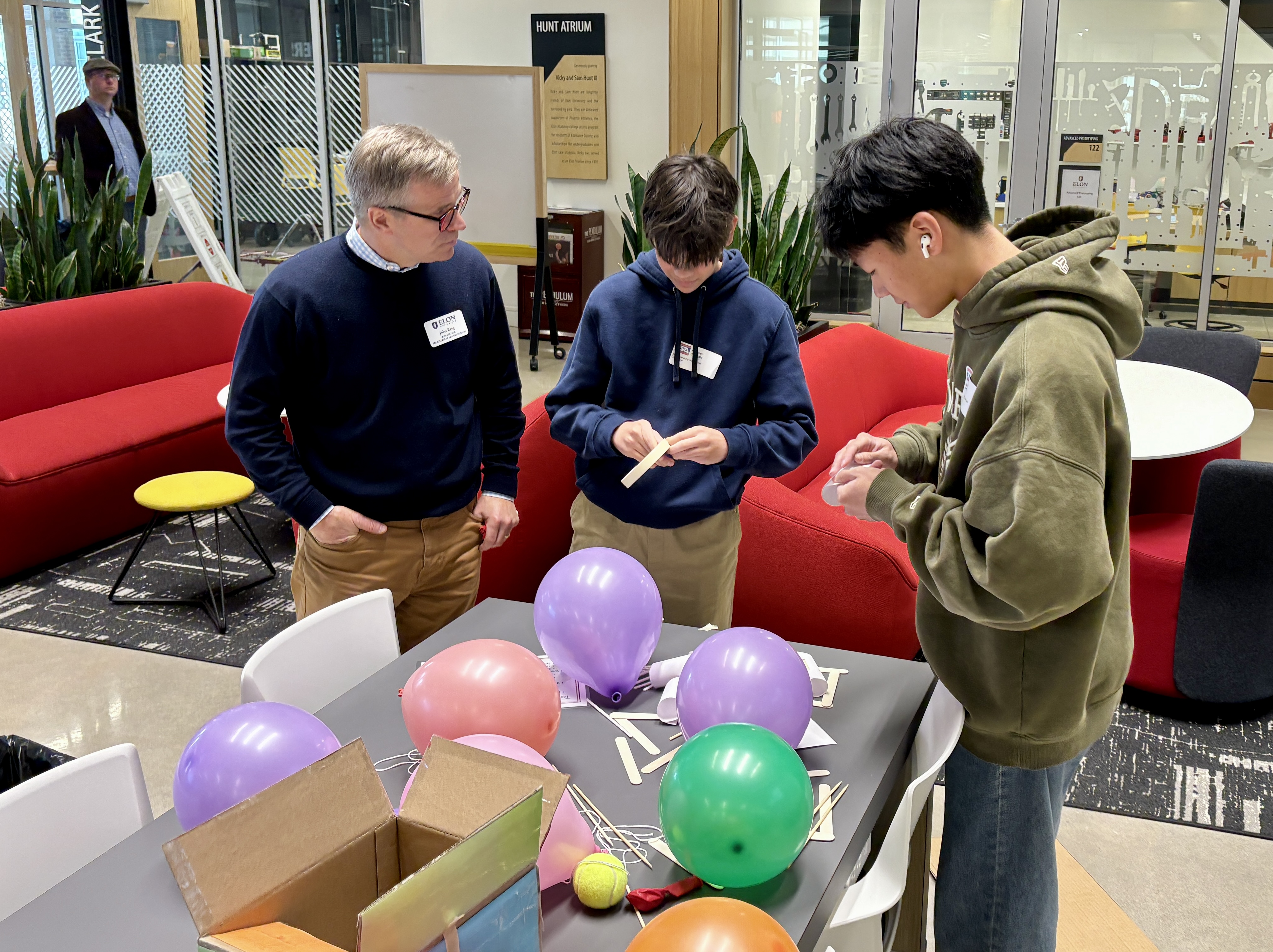 Two male high school students work with adhesive tape and popsicle sticks on a table covered with balloons. A member of male staff speaks to them while they work.