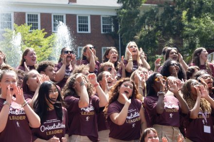 A large group of students wearing matching maroon "Orientation Leader" shirts, enthusiastically cheering outdoors near a fountain with a brick building and trees in the background.