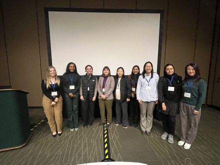 Group photo of nine professionals in business attire, standing in front of a presentation screen at a conference. Each individual is wearing a name badge.