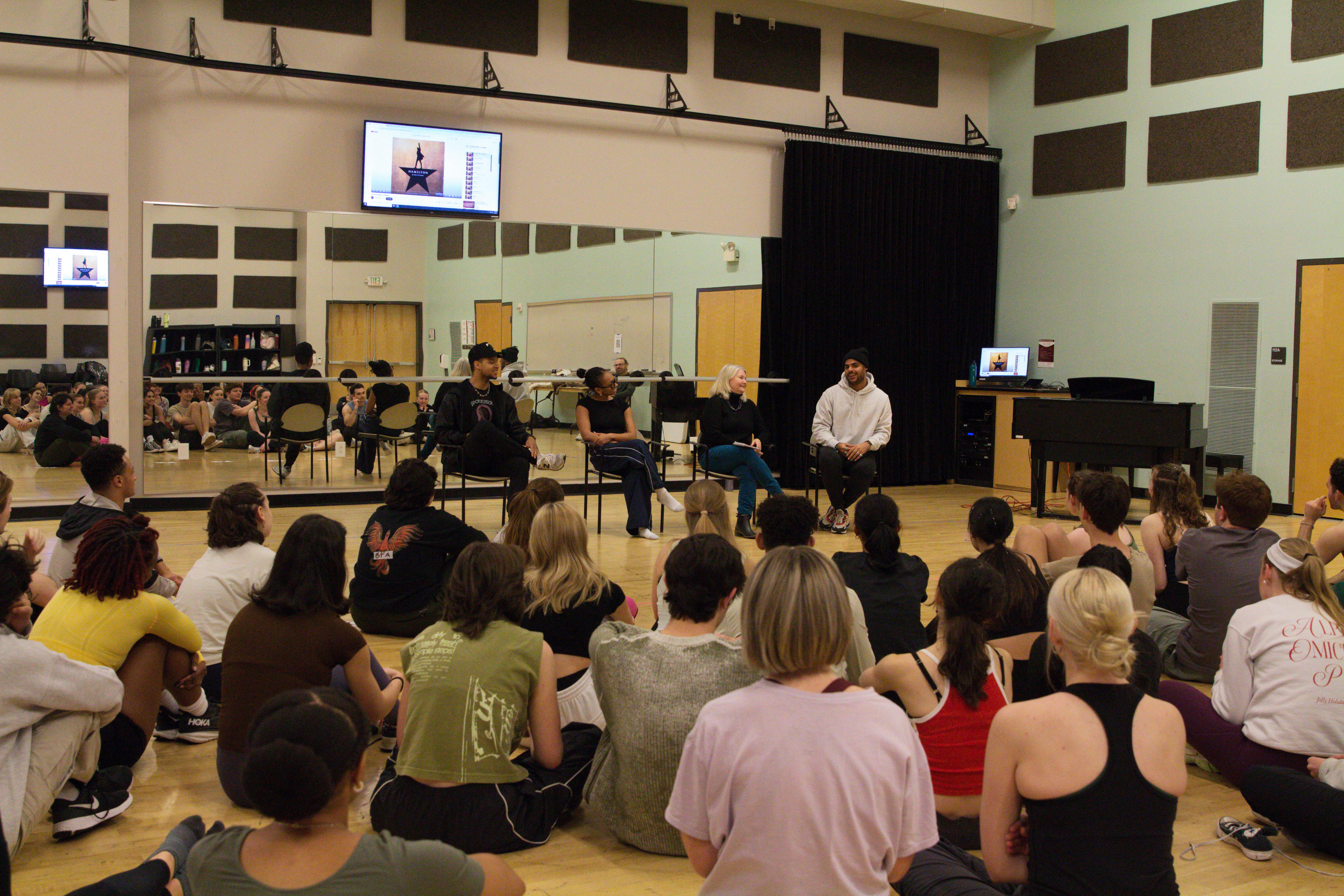 a large group of students seated on the floor in front of a row of chairs in a dance rehearsal studio.