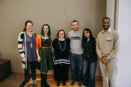 Six people stand for a photo in front of a beige wall