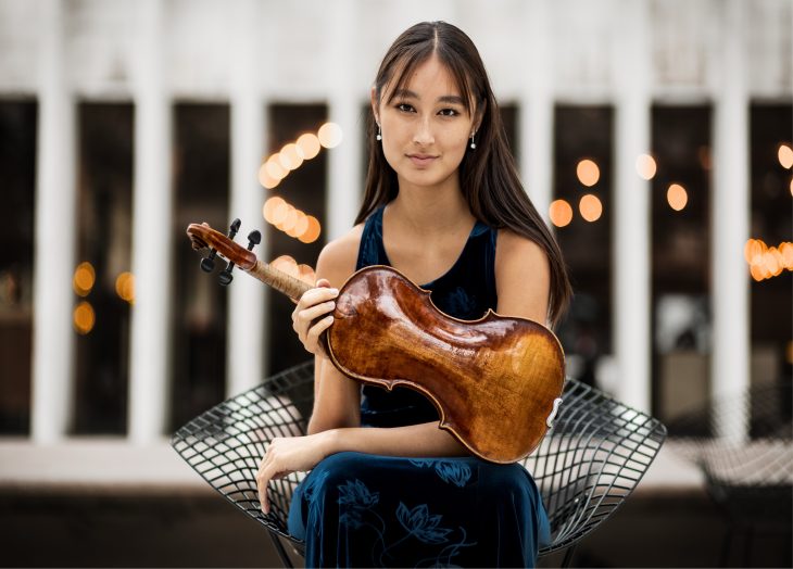 A woman holding a violin and looking into the camera. She has dark hair and is wearing a blue dress, sitting in front of a building with interesting architectural features.