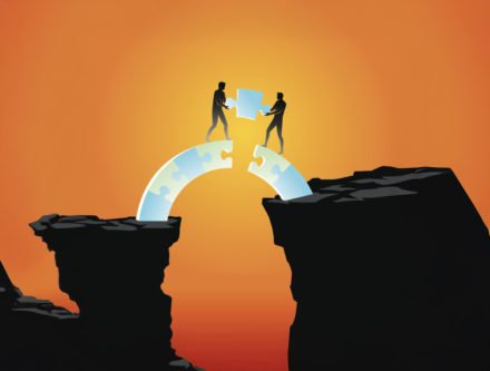 Two silhouetted figures connect puzzle pieces to bridge a gap between cliffs, with a sunset in the background.