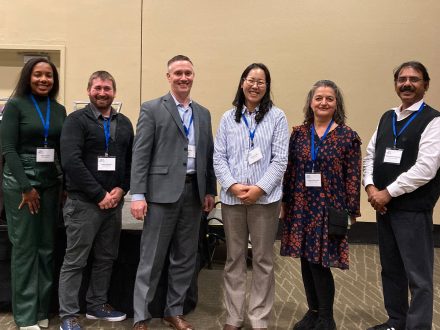 Group of six professionals, wearing conference name tags, smiling and standing together at an indoor event.