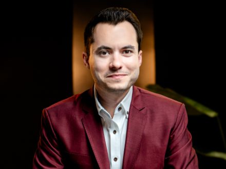 Man in maroon blazer and gray shirt stares directly at the camera