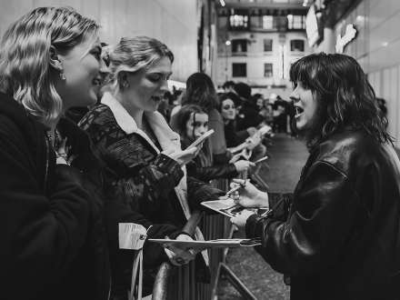 This black and white photograph captures a lively moment at a stage door or fan event. A woman wearing a leather jacket, likely a performer, is enthusiastically signing autographs and engaging with fans. Her expressive face and open mouth suggest she is in the middle of an animated conversation. Two young women in the foreground, both smiling, hold out programs or photos for signing. One of them, wearing a warm coat with a shearling collar, is holding a phone as if taking a picture or recording the moment. The other, in a dark jacket, appears delighted, with her hand on her chest. A crowd of fans extends into the background, forming a queue along metal barricades, all eagerly waiting for their chance to meet the performer. The setting appears to be outside a theater, with bright lights reflecting off modern building panels and classic architecture in the distance. The image conveys excitement, connection, and the joy of meeting someone admired.