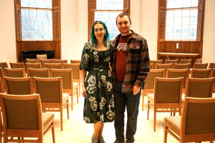 Two people stand in a room with windows behind them and chairs