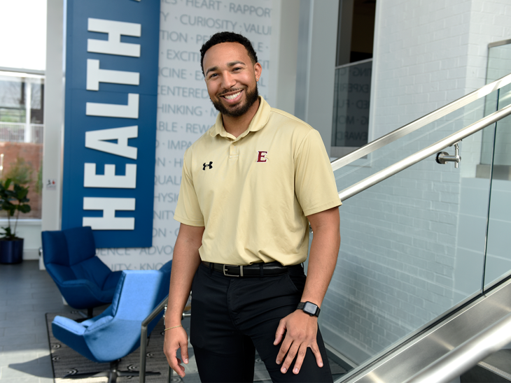 This image features a young man with a well-groomed beard and short curly hair, standing in a modern indoor setting. He is smiling warmly and looking directly at the camera. He is wearing a light yellow polo shirt with an "E" logo embroidered on the left side of his chest, along with an Under Armour logo on the right side. He pairs the shirt with black pants, a black belt, and a smartwatch on his left wrist. The background showcases a professional and stylish interior with a large blue sign that prominently displays the word "HEALTH" in white capital letters. The walls are decorated with motivational words in a subtle, modern font. A glass railing is visible on the right, and a set of comfortable blue chairs with a sleek design is positioned in the lounge area. There are also indoor plants adding a touch of greenery. The overall atmosphere suggests a health, wellness, or academic setting, possibly related to a university or health facility. The subject’s confident and approachable demeanor aligns with a professional or educational role.