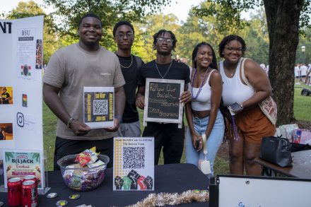 This photo captures a group of five smiling college students at what appears to be a campus organization fair. They’re standing behind a display table representing the Black Student Union (BSU). Here are some key details: The students are posing together outdoors with trees and other fair attendees in the background. Two of the students are holding signs—one has a QR code with text saying “JOIN BSU” and “Scan Now,” while another has a small chalkboard sign reading: “Follow Our Socials…” Instagram: @elonbsu TikTok: @elonbsu There’s a large bowl of candy and beaded necklaces on the table, suggesting giveaways to attract people. A trifold board to the left has the title "BSU" with info on what the organization does, images, and social media links. A flyer at the front of the table advertises a BSU event called the B(l)ack Cookout. Everyone in the group looks happy and engaged, showing off a welcoming and inclusive vibe.