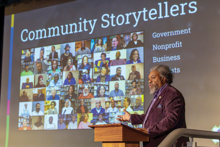 A man stands behind a podium gesturing toward a screen. The screen features a mosaic of photos of different people and reads "Community Storytellers"