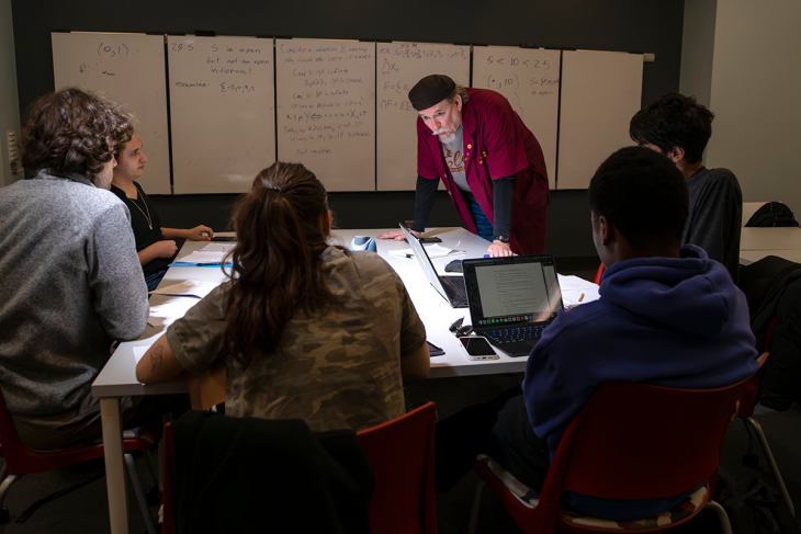 A math professor stands and leans against a desk and talks to a group of students who are seated around the desk. A white board with mathematical equations written on it is on the wall behind him.