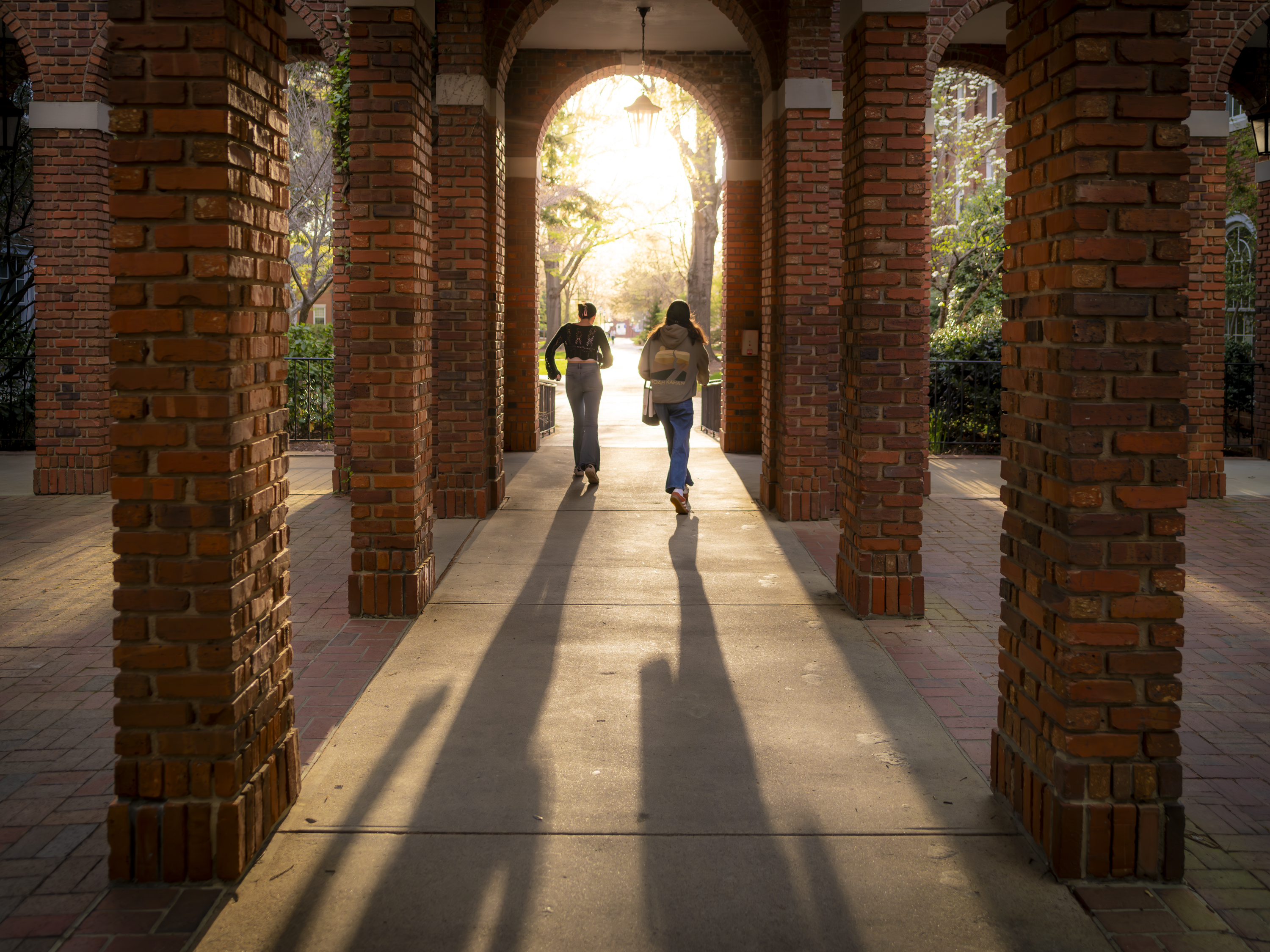Two individuals walking through a brick archway, bathed in sunlight with long shadows stretching ahead.
