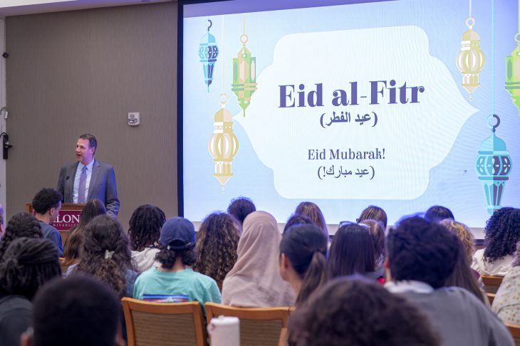 A person stands at a podium addressing an audience during an Eid al-Fitr celebration. The presentation screen behind displays "Eid al-Fitr" and "Eid Mubarak!" in both English and Arabic script.