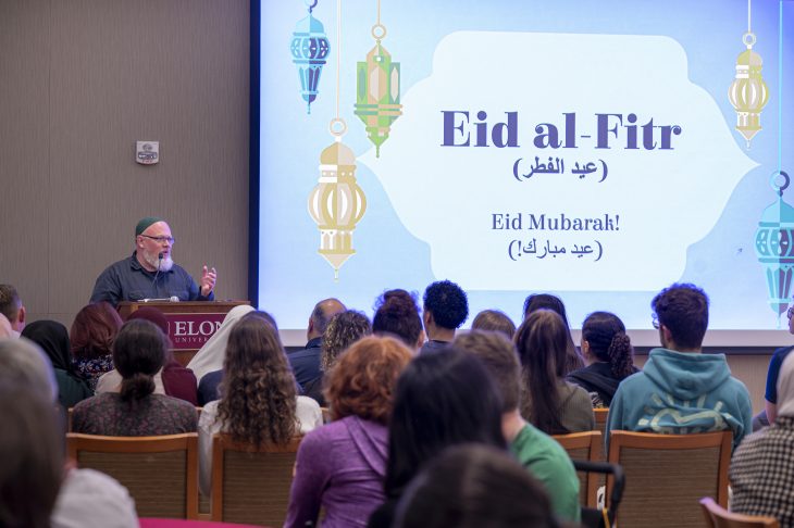 Speaker at a podium discussing Eid al-Fitr with an audience in a conference room, with decorative lanterns and "Eid Mubarak" text displayed on a presentation screen.