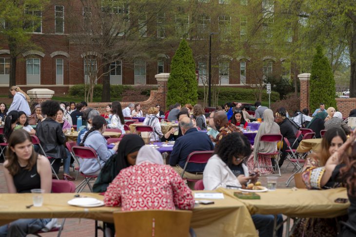 Outdoor gathering at a university with people seated at tables covered with yellow tablecloths, dining and conversing. Classic brick buildings and greenery surround the area.