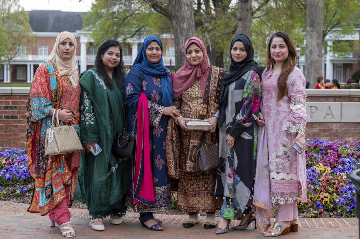 A group of six individuals in traditional South Asian attire, standing together and smiling in a garden with colorful flowers.