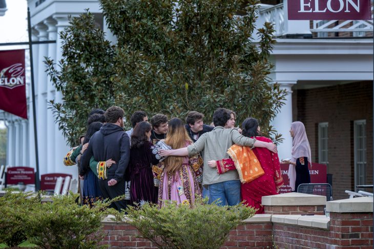 Group of people in diverse attire huddled together on a university campus with an "Elon" banner in the background.