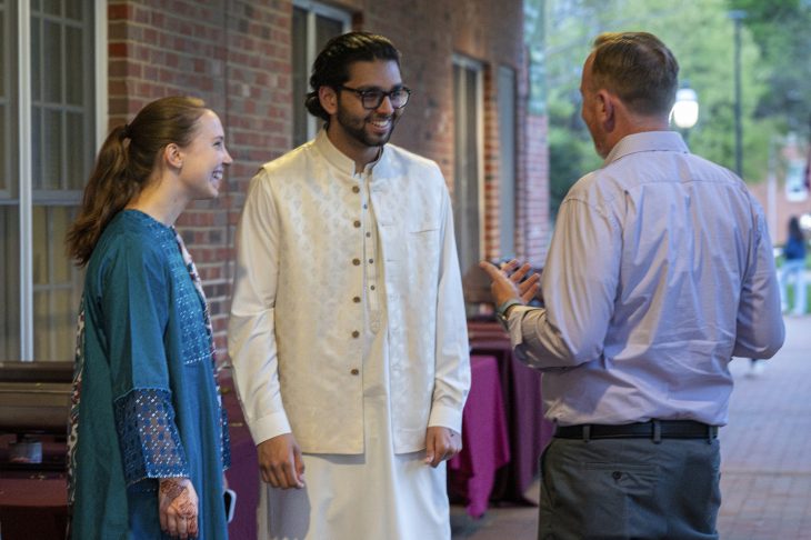 Three people engaging in a friendly conversation outdoors, one in a traditional South Asian attire and the other two in western casual clothing.