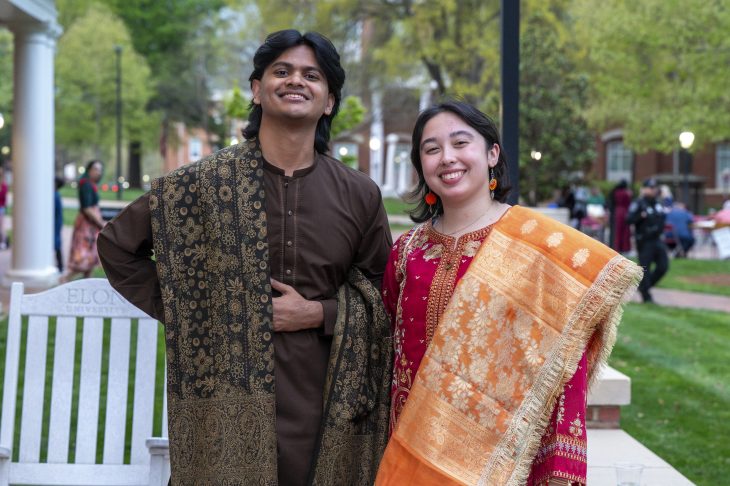 Two people wearing traditional South Asian attire are standing together and smiling in a campus setting at Elon University. One is dressed in a brown and gold kurta, and the other in a red saree with an orange border.