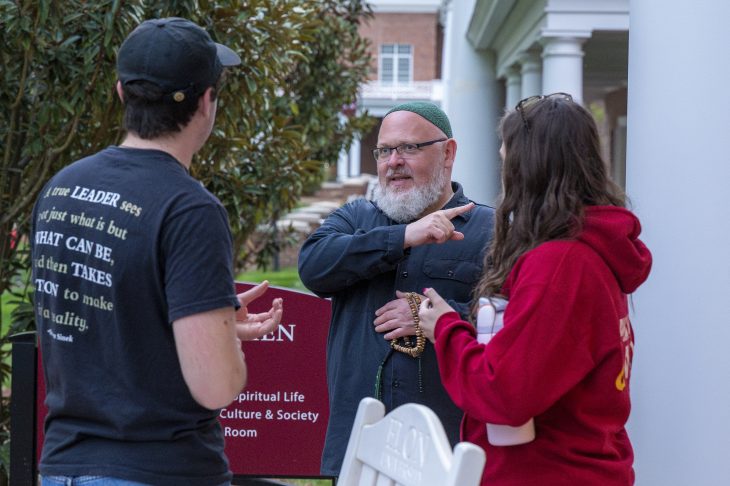 Three individuals are engaged in a conversation outside a building. One person is pointing and speaking animatedly.