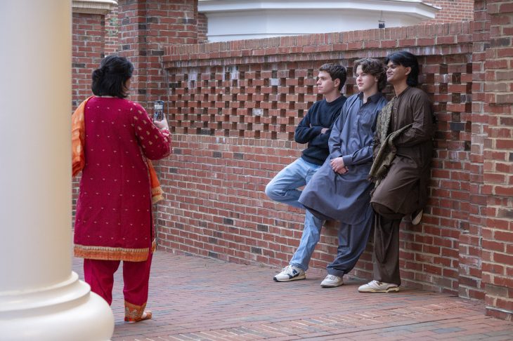 A person in a red traditional outfit is speaking to three individuals seated on a brick wall.