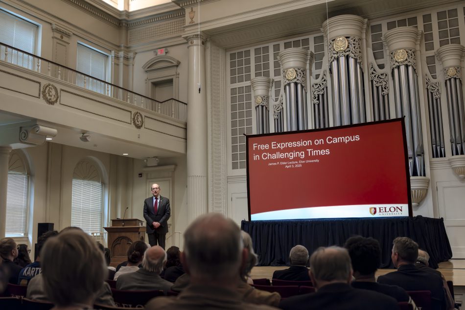 Generate by AI Assist Presentation titled 'Free Expression on Campus in Challenging Times' at an Elon University event, featuring a speaker standing next to a large screen in an auditorium filled with attendees.