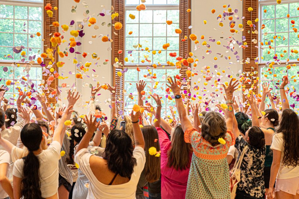 Group of people joyfully tossing colorful flower petals into the air in a room with large windows.