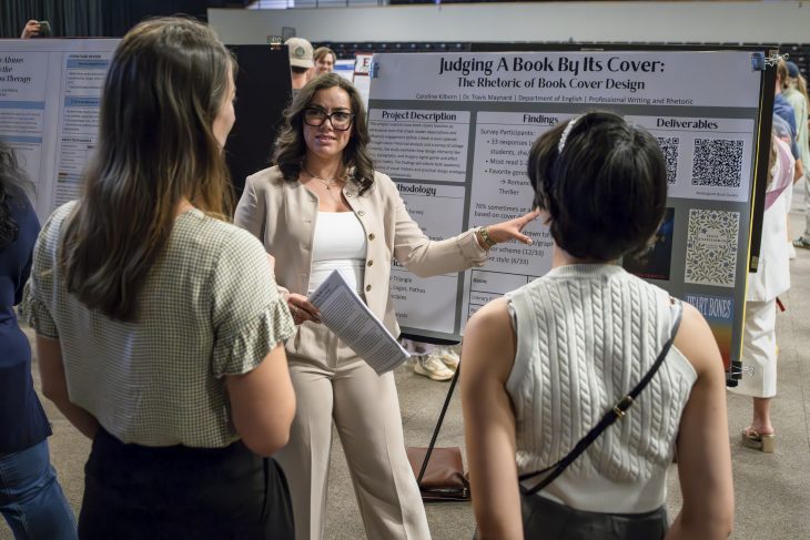 A young woman presents a large poster presentation to two people