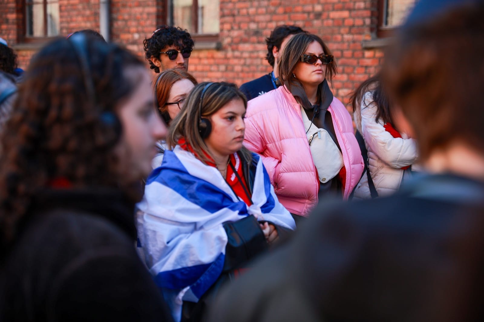 A group of people wearing headphones listens attentively during an outdoor tour. One woman in the center is draped in a flag, while others around her wear warm clothing and appear focused.