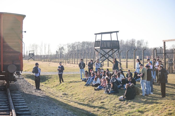 Visitors gather for a guided educational tour at Auschwitz-Birkenau, with the railway and watchtower in view under clear skies.