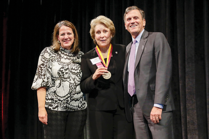 Elon University President Connie Ledoux Book, alumna Debbie Yow-Bowden and Trustee Ed Moriarty pose for a photo onstage. Debbie wears a medallion around her neck with a maroon and gold ribbon.
