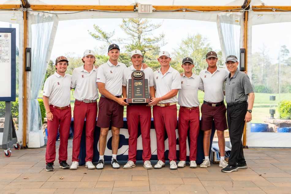 A group of Elon men's golf team members posing outdoors with a championship trophy. They are dressed in matching maroon and white uniforms.