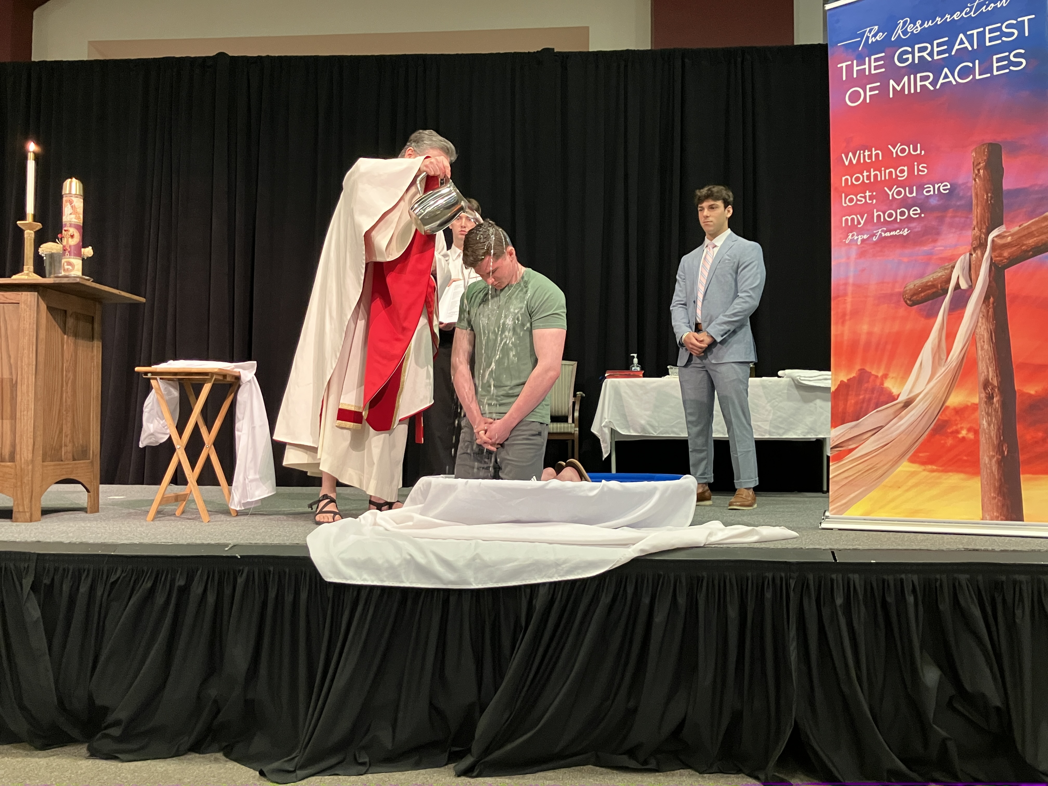 priest pours water on student kneeling in a basin as another student stands behind.