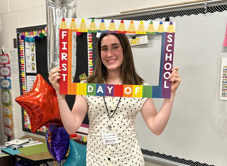 Riley Fennelle, an Ƶ student-teacher, stands in front of a white board in an elementary school classroom. She holds a cardboard frame with a pattern of rainbow pencils that says "first day of school" around her face.