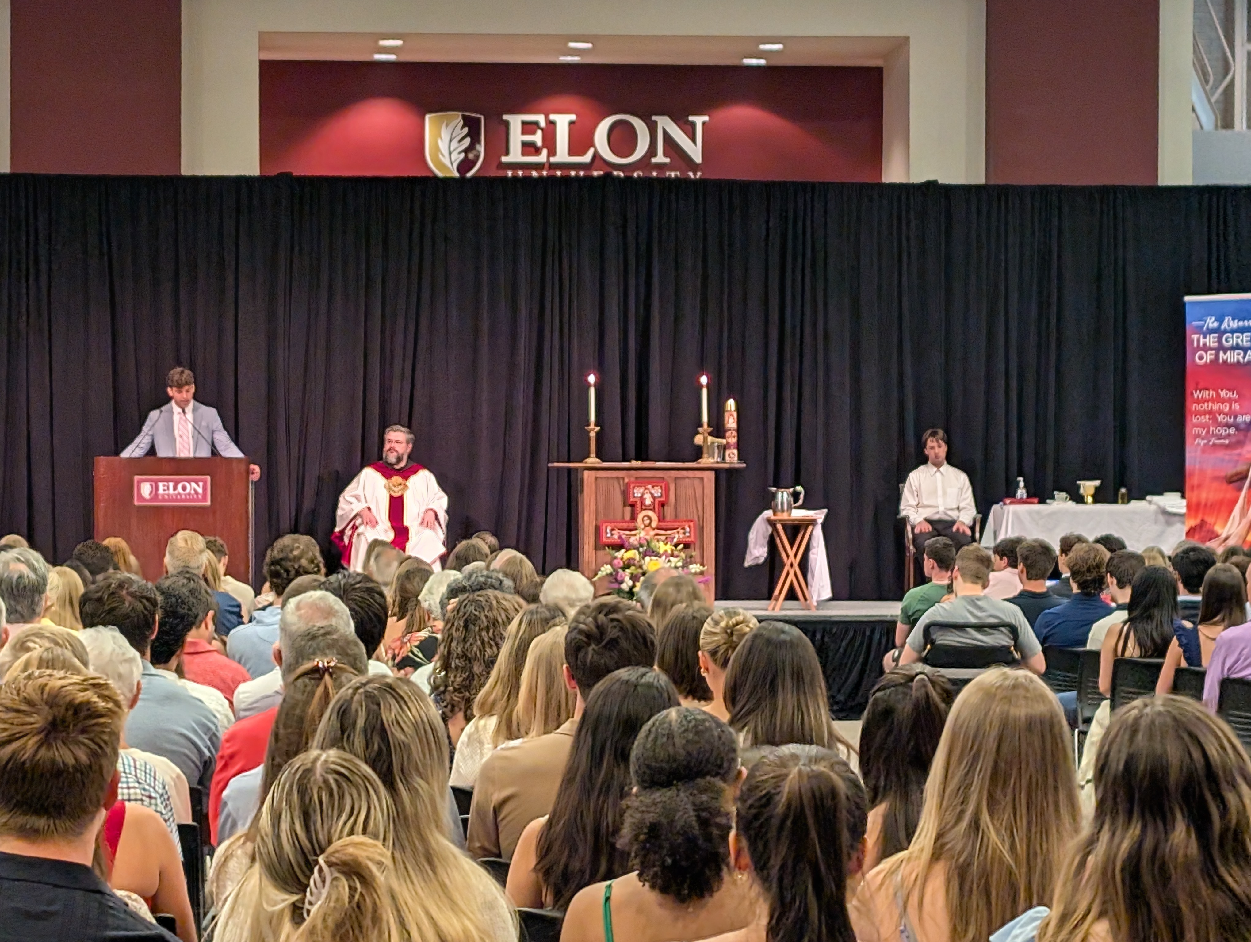 Student at podium with priest and altar server sitting on stage with altar in the middle.