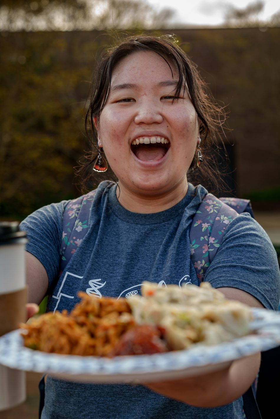A person holding a plate of food and smiling broadly, with a tranquil outdoor setting in the background.