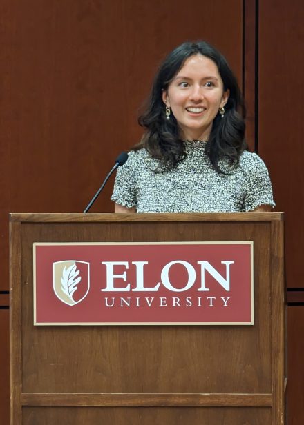 A photo of Alana Evora '23 in a green dress, speaking at a podium with the Elon University logo on it.