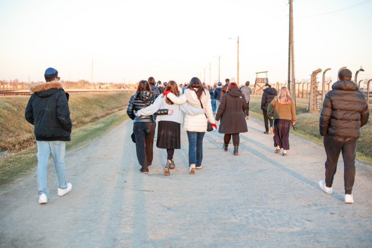 A group of people walks down a dirt path, some with arms around each other for support. The setting appears solemn, with barbed wire fences and guard towers visible along the sides of the path.