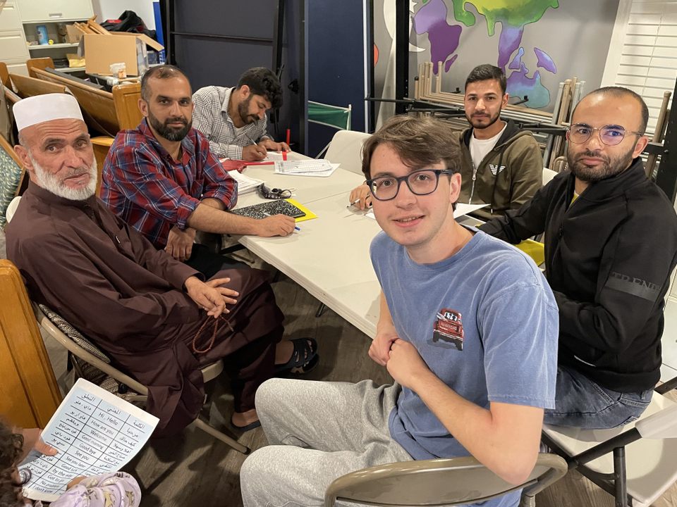 A group of men, including a student in an Elon T-shirt, sit around a table with papers and worksheets, engaged in what appears to be an English language or literacy lesson in a welcoming community space.