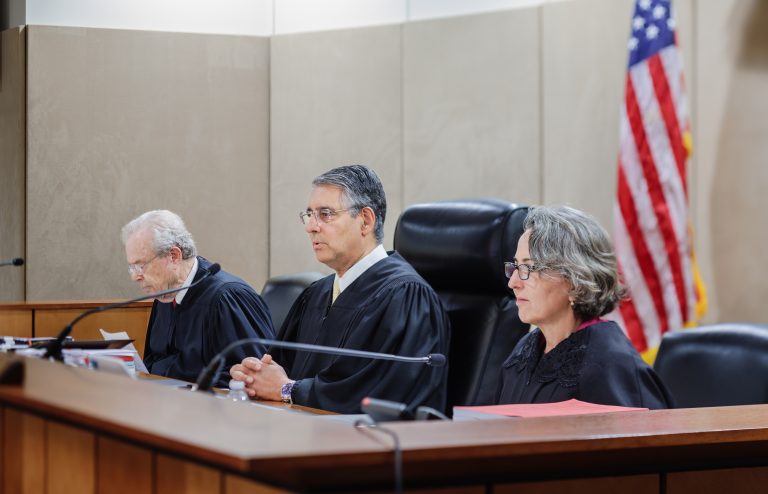 Three judges, two male and one female, sit at the dais in a courtroom. They are wearing black robes. U.S. and North Carolina Flags are seen behind them.