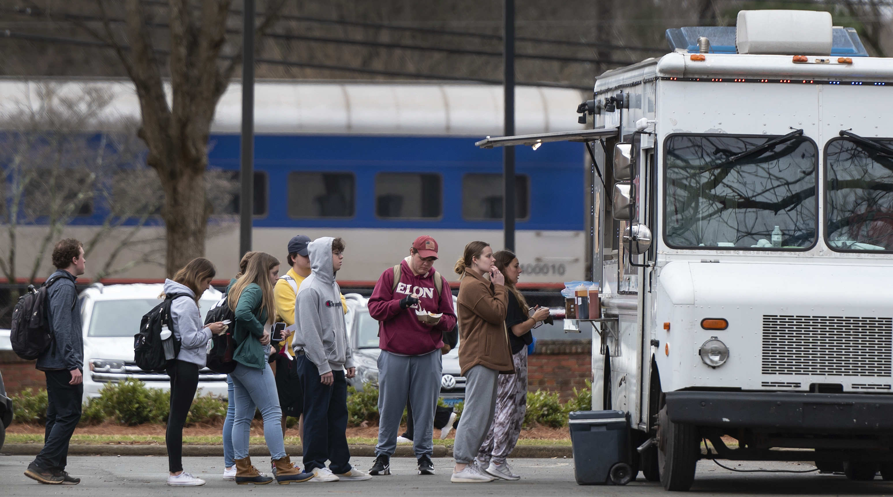 A group of people stands in line at a white food truck parked outdoors, with several holding food or looking at their phones. Most are casually dressed, wearing hoodies, jackets, or sweatshirts, and backpacks. In the background, a blue and white passenger train passes by, partially obscured by trees and utility poles.