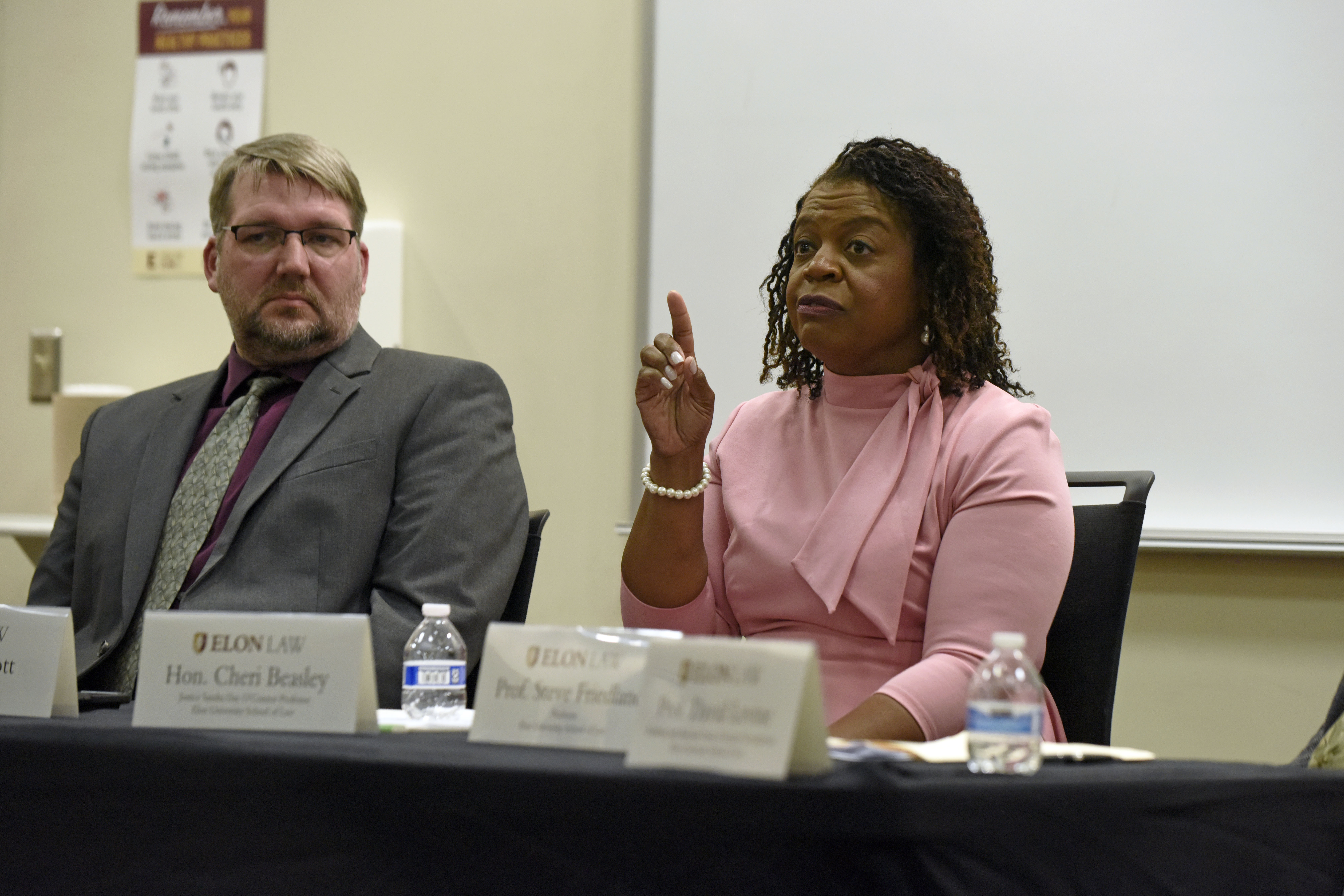 A woman wearing a pink sweater gestures pointing her finger in the air as she speaks. A man to her left listens intently.