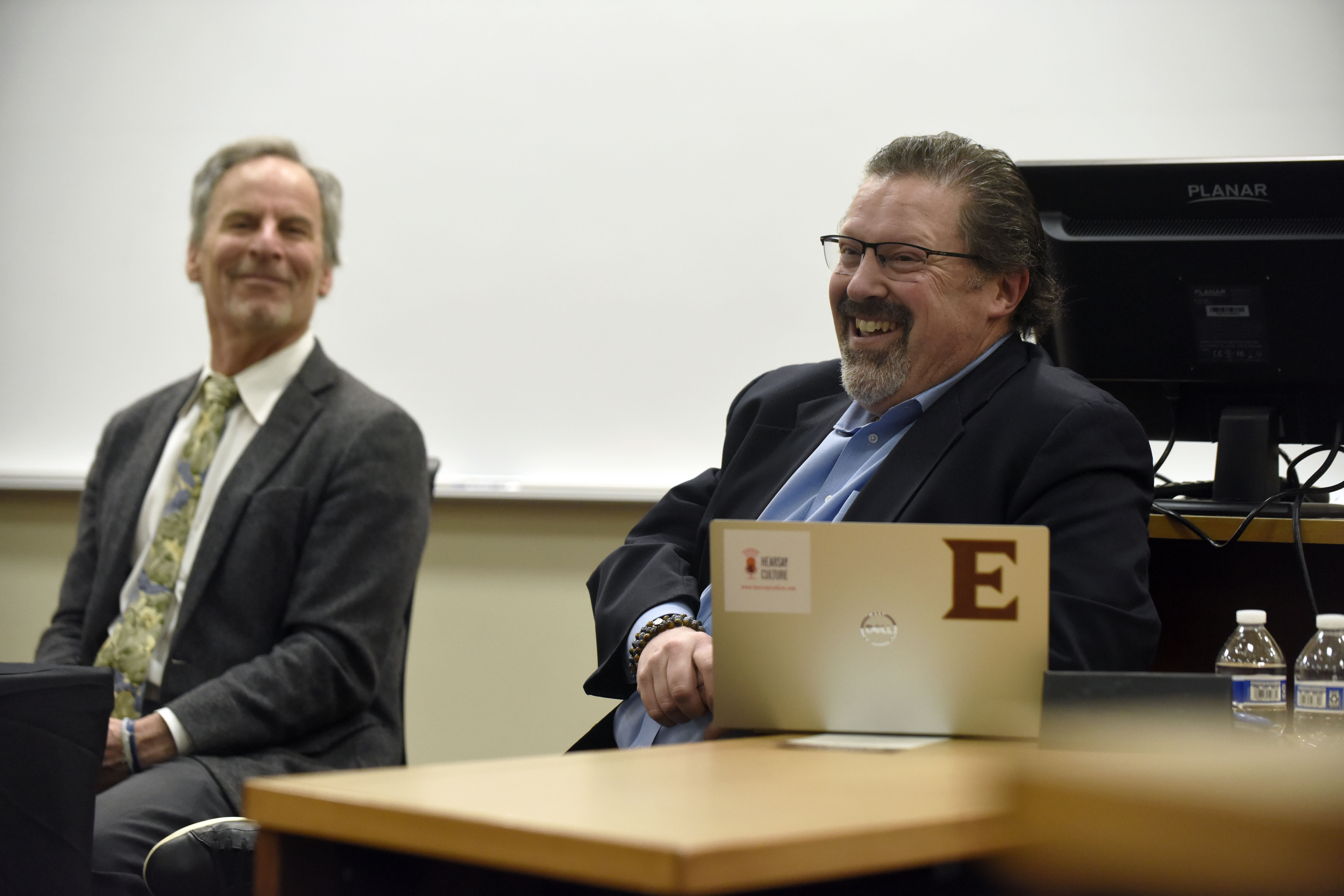 Two men seen smiling and laughing. One has a laptop open with a large monogram E on it.