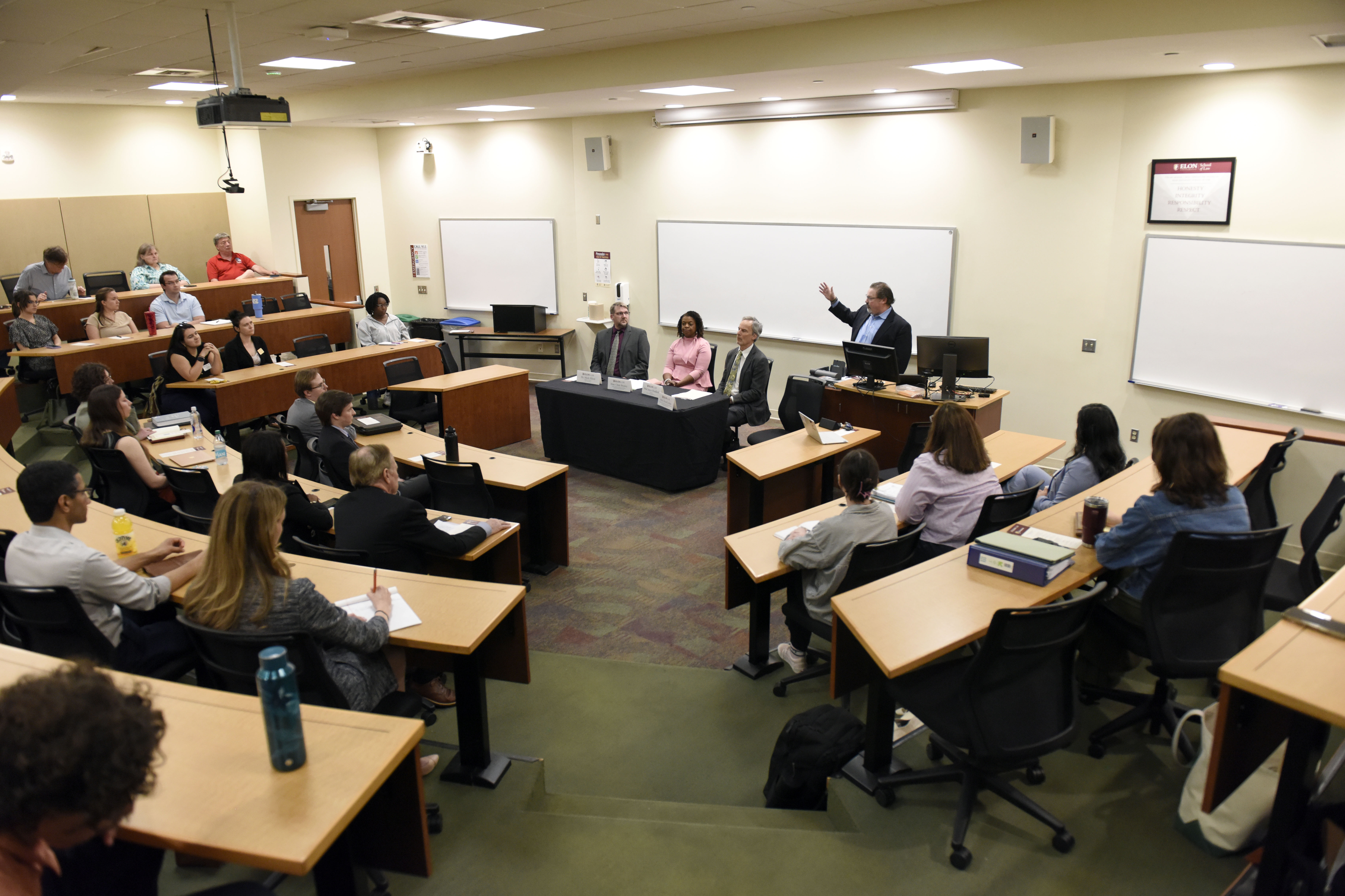 A large amphitheatre style classroom with a panel of three speakers and a man at a podium at the front of the room.The room is filled with people listening to remarks.