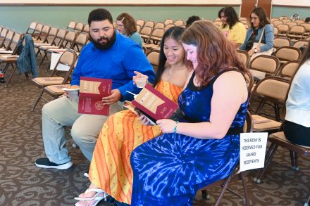 Three people sit together in a row of chairs, smiling and reading from maroon and gold event programs labeled “Elon Leadership Awards Ceremony 2025.” They appear to be award recipients, as a sign on their row reads “This row is reserved for award recipients.” The person on the left, wearing a blue shirt, holds a cup and plate; the two on the right wear colorful dresses. Other attendees are seated in the background, and many chairs remain empty in the event space.