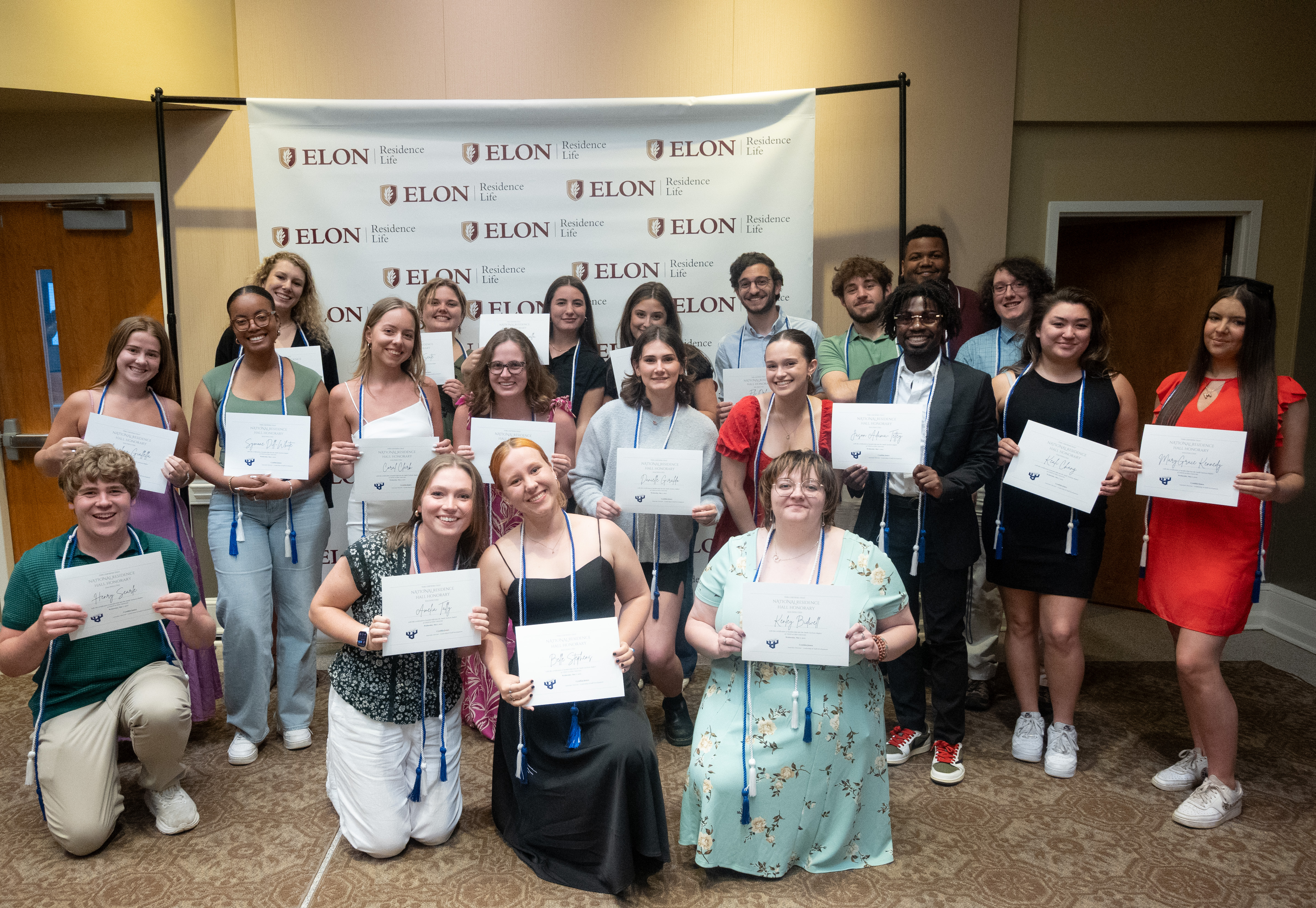 A large group of smiling students pose in front of an Elon Residence Life backdrop, proudly holding certificates and wearing honor cords.